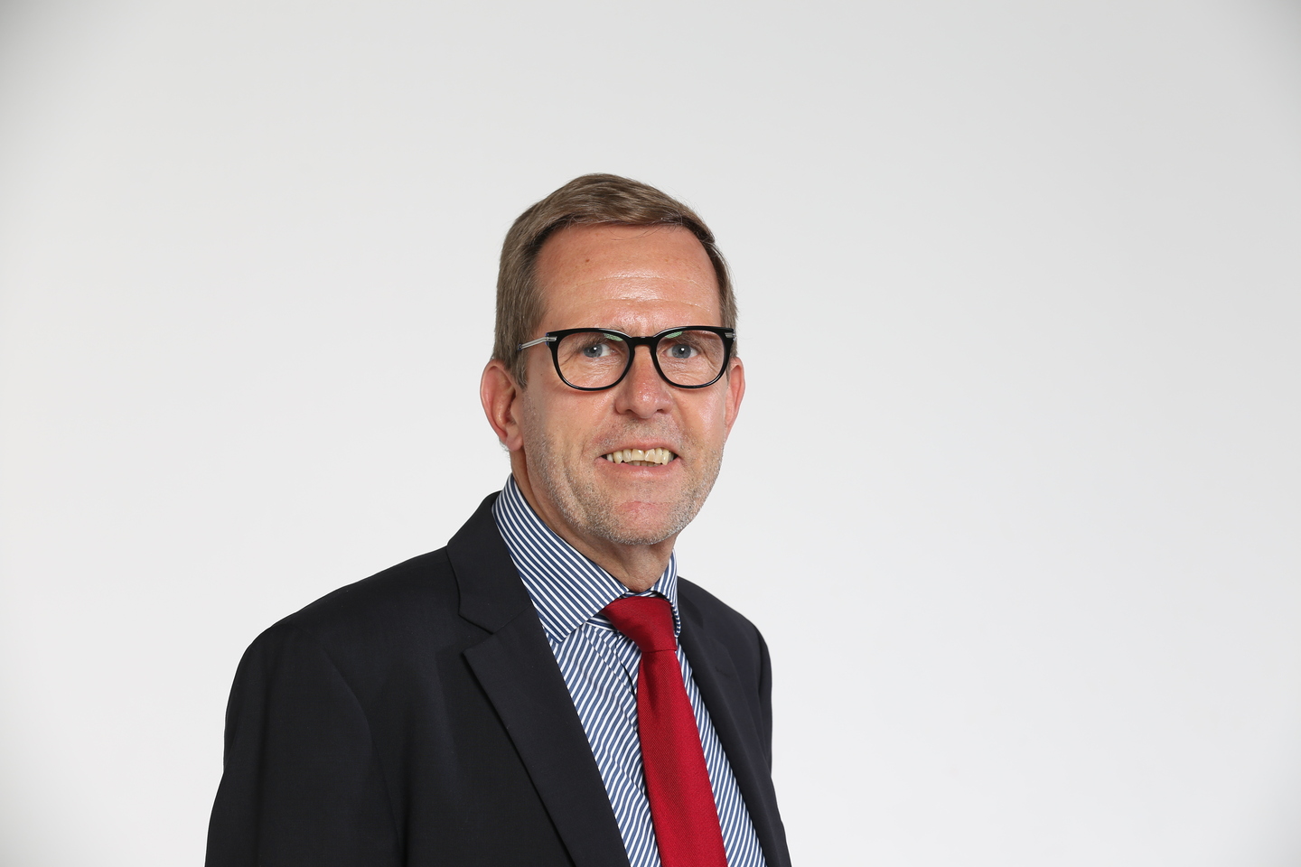 Ein Mann mit Brille, Hemd und roter Krawatte lächelt vor einem weißen Hintergrund.